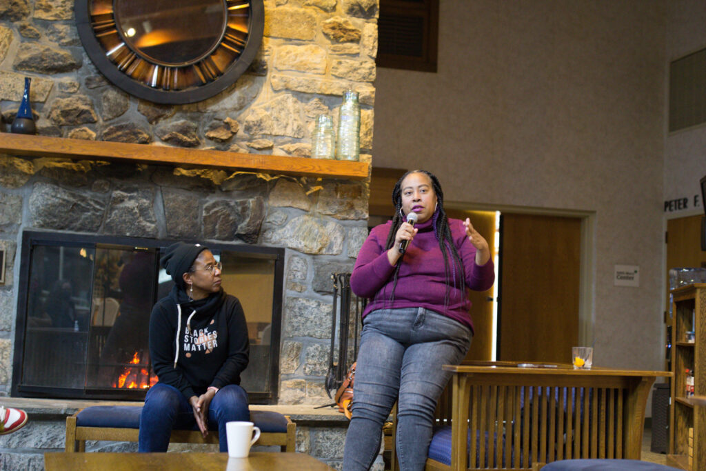 At the main lounge of the Edith Macy Center, Angela Tucker sits at the arm of a chair speaking into a microphone. Leslie Fields-Cruz looks up at her while sitting nearby the fireplace.