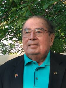 Headshot of a Native American man wearing wire glasses, an aqua-toned poloshirt and a dark blazer posed in front of a tree