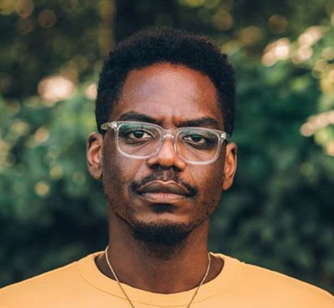 a mustached black man wearing clear glasses and a high-top fade hairstyle stares straight ahead wearing a gold-colored t-shirt