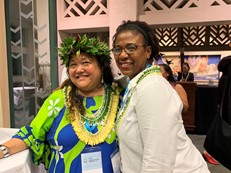 a smiling Hawaiian woman in a blue and green colorful top wearing a yellow lei and a leafy headband stands beside a black woman in a cream colored blazer wearing a green lei