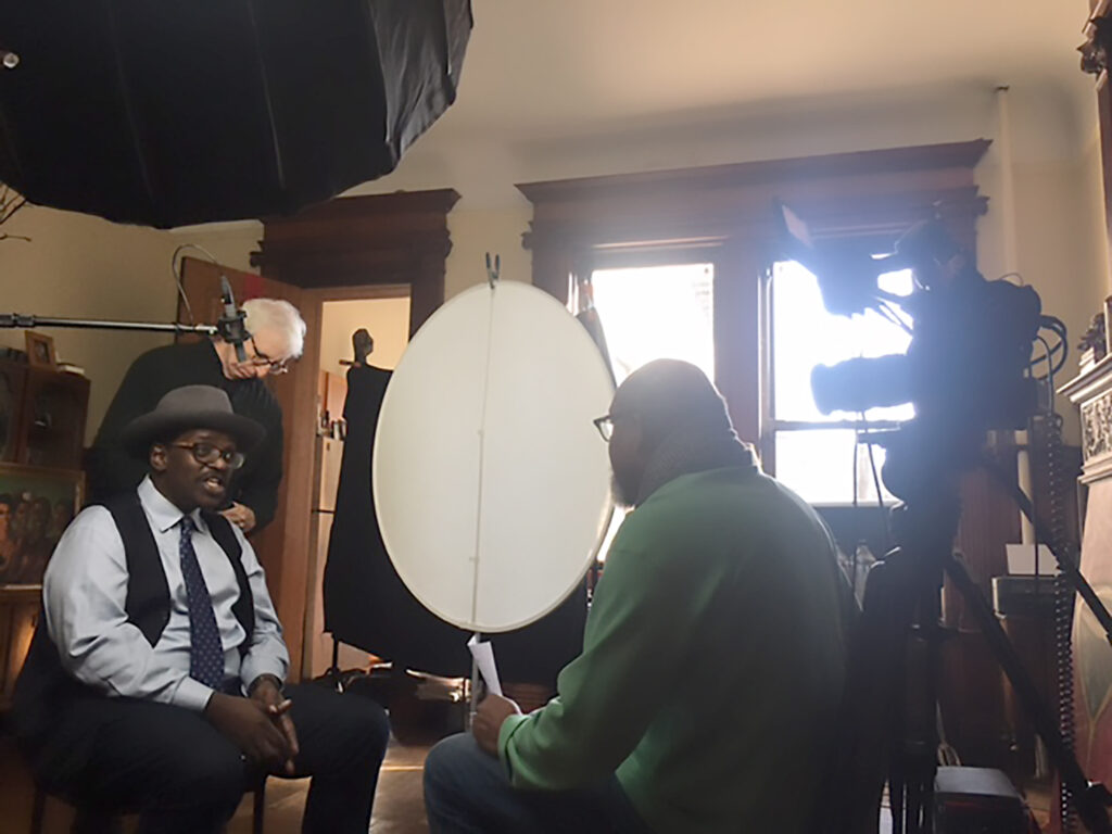 two black men sit face to face in an interview with light, camera, and audio equipment in the background and a white man stands in the background staring down at a piece of equipment that is obscured from view.