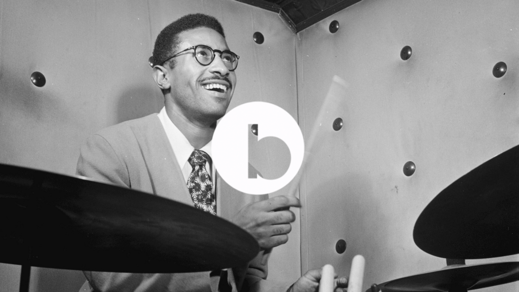 lack man wearing dark-rimmed glasses joyfully playing a drum kit in a sound studio