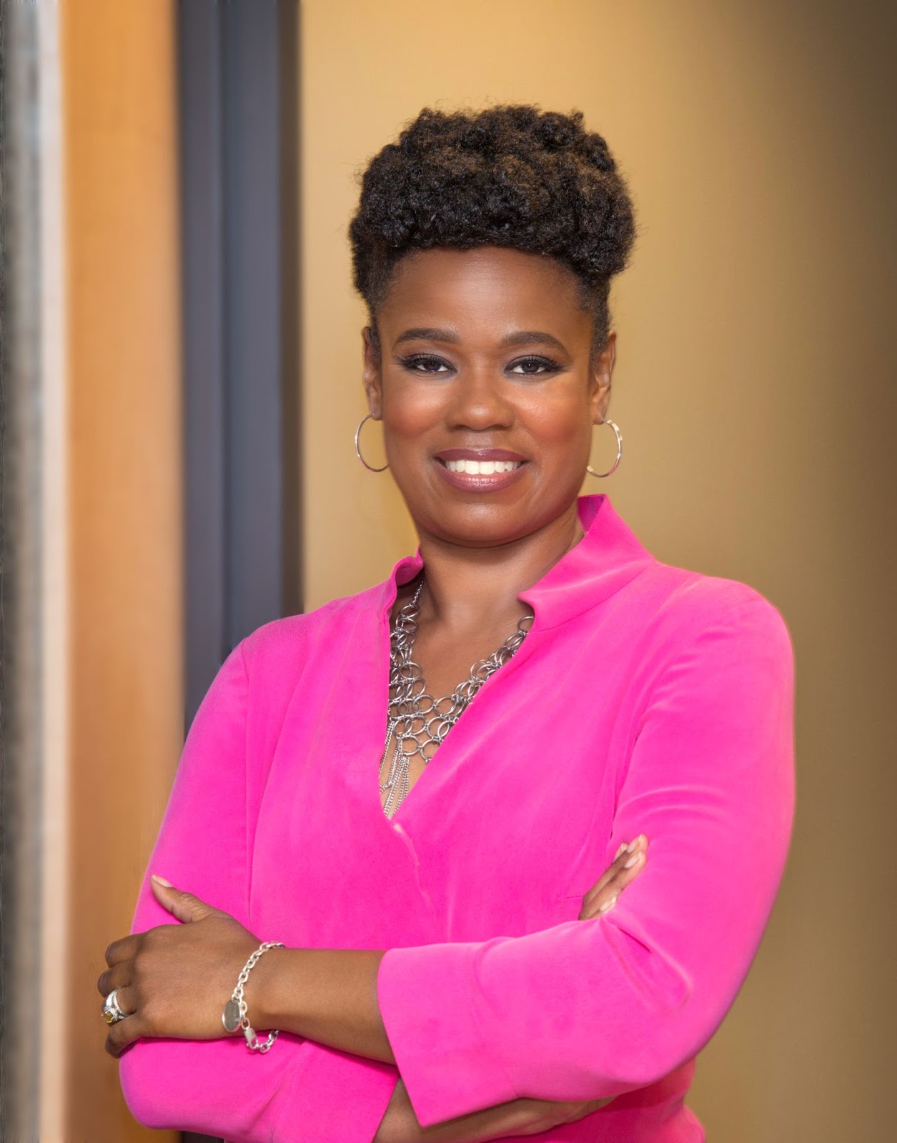 Smiling black woman with arms folded across her chest, hair neatly woven into a bun on top of her head, wearing a fuchsia blouse.