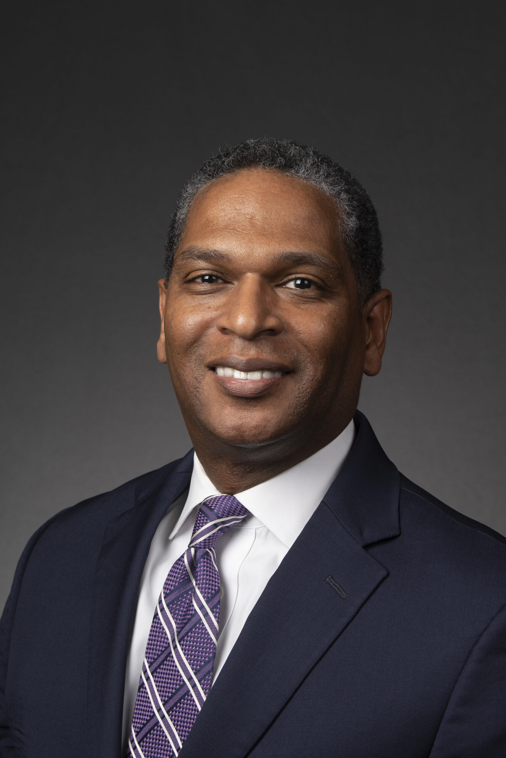 Smiling black man with mixed-gray hair wearing a dark suit, white shirt and purple, black and white patterned tie