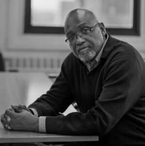 black man in dark sweater looking at camera to his left while seated at desk in a classroom