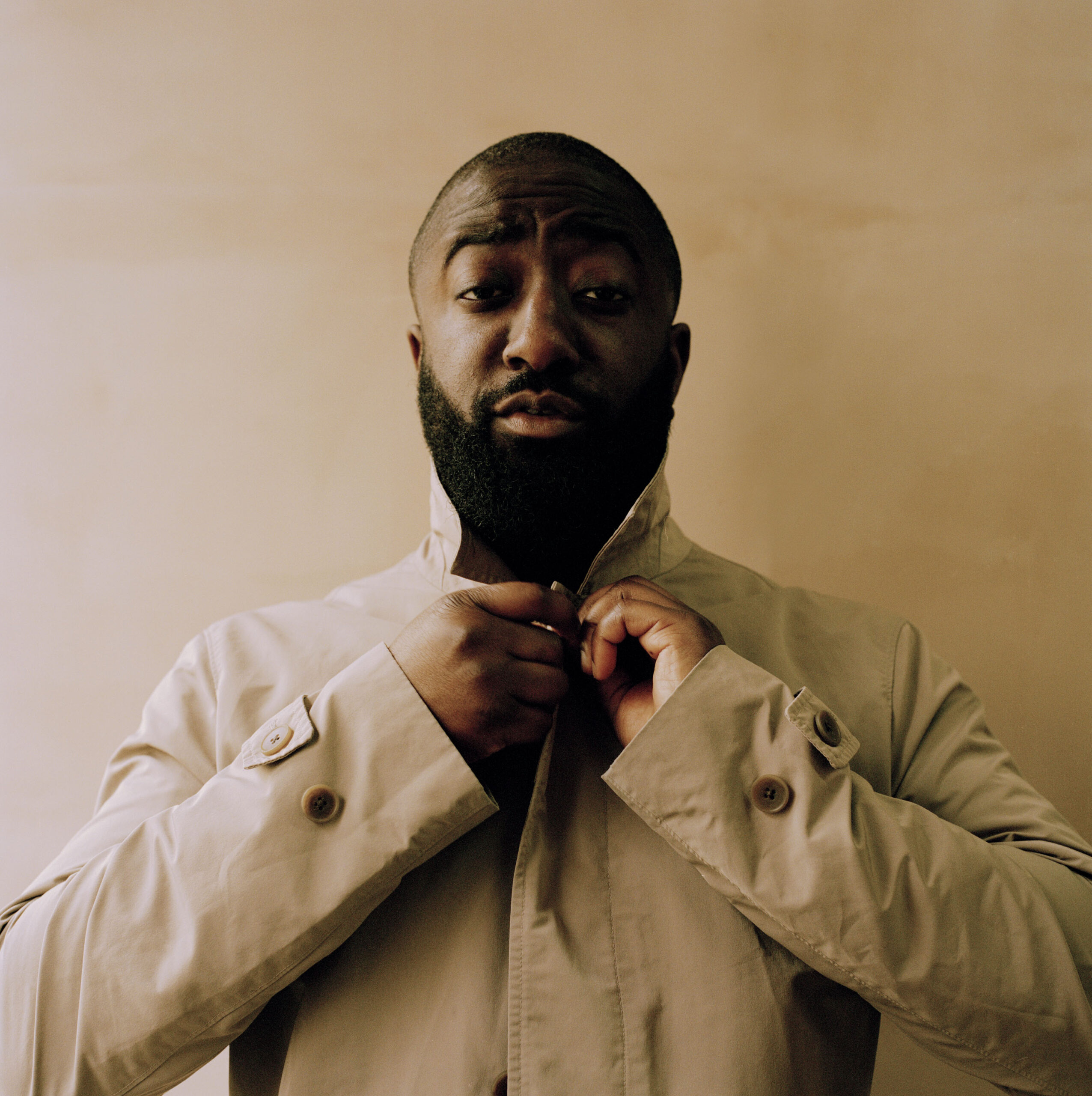 bearded black man in beige shirt looks at camera while buttoning top botton