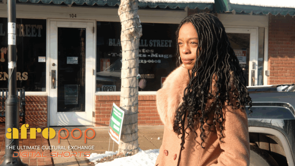 woman with long dark curly hair in a shearling coat faces frame left in front of a storefront