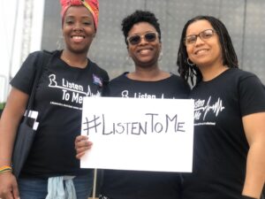 three women in black branded t-shirts holding a white poster with the hashtag for the film #listen to me