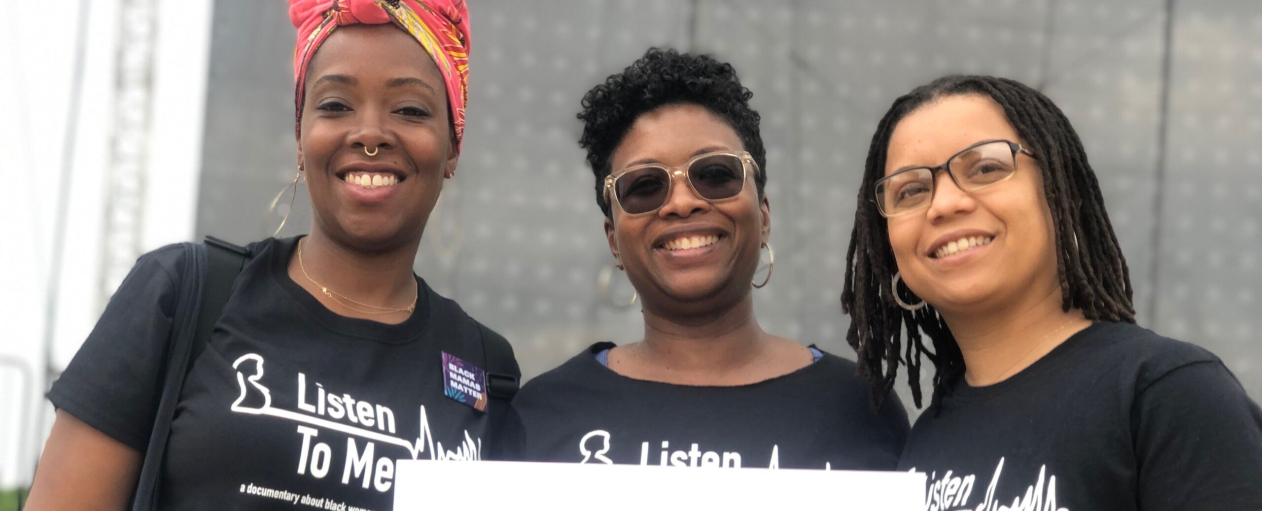 three women in black branded t-shirts holding a white poster with the hashtag for the film #listen to me
